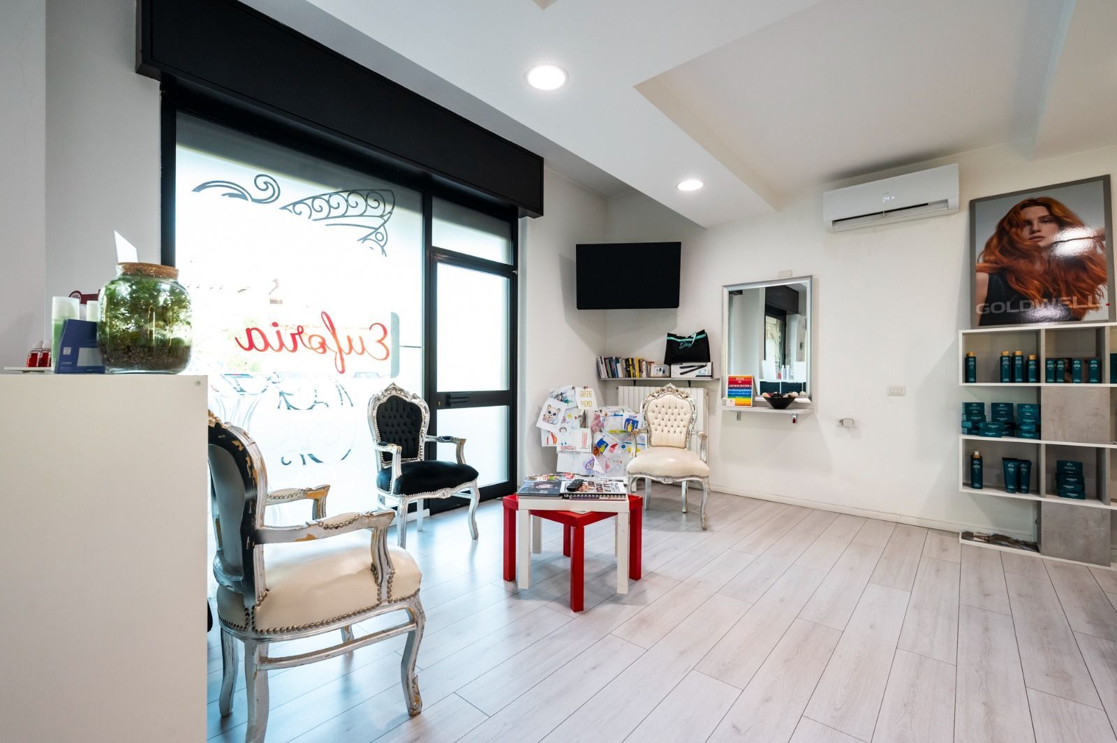 Interno di un salone da parrucchiere: pareti bianche, pavimento in legno, sedie decorate, tavolino rosso, grande finestra, prodotti sugli scaffali.