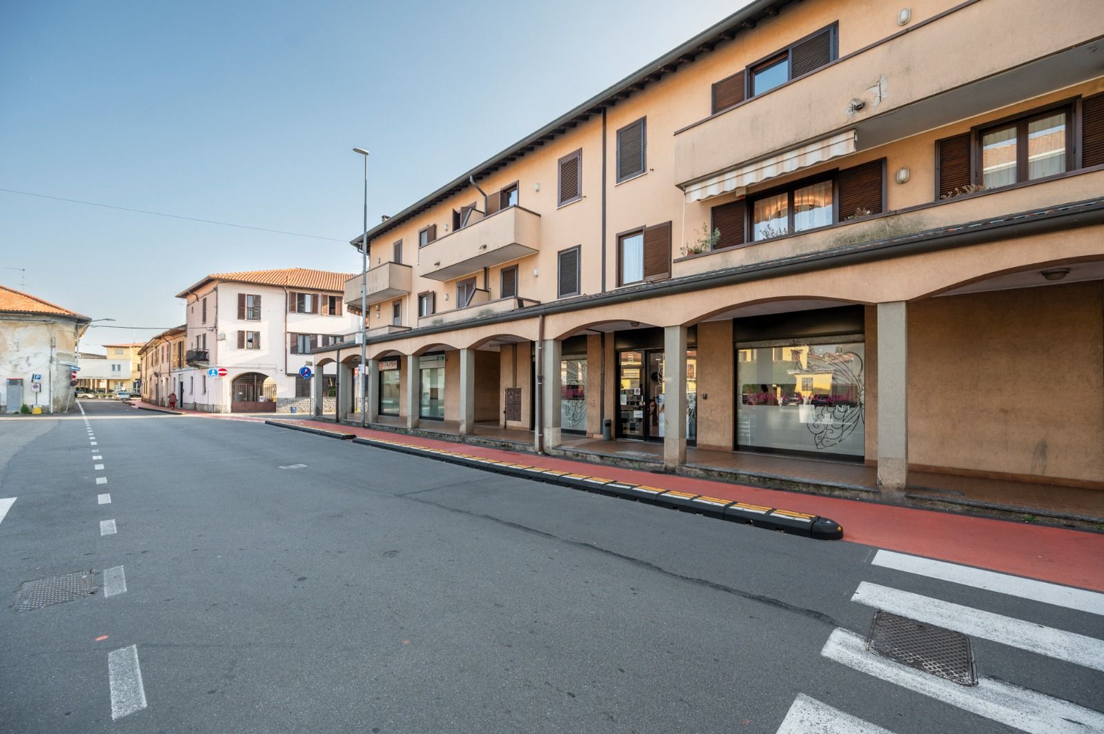 Veduta di edifici beige con negozi, tende da sole e balconi. Attraversamento pedonale in primo piano.