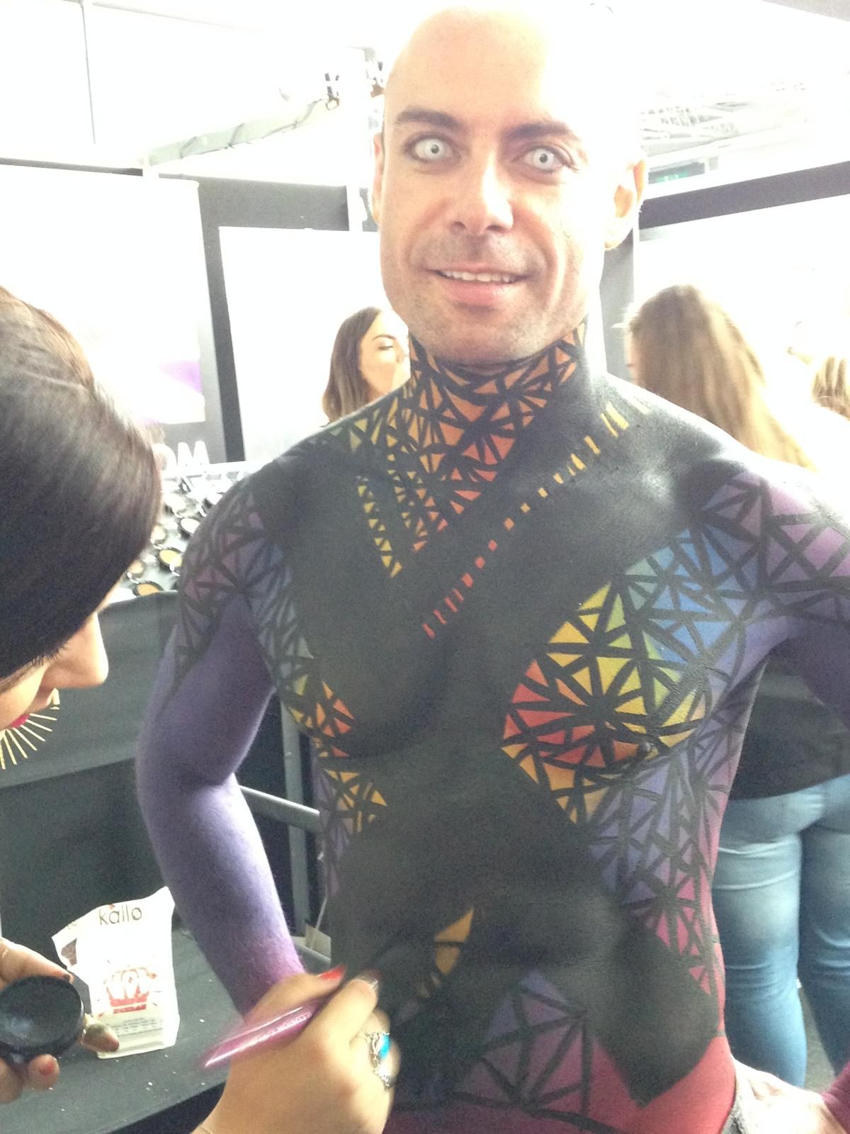 Uomo con body painting, motivi geometrici colorati, occhi bianchi. Viene dipinto.