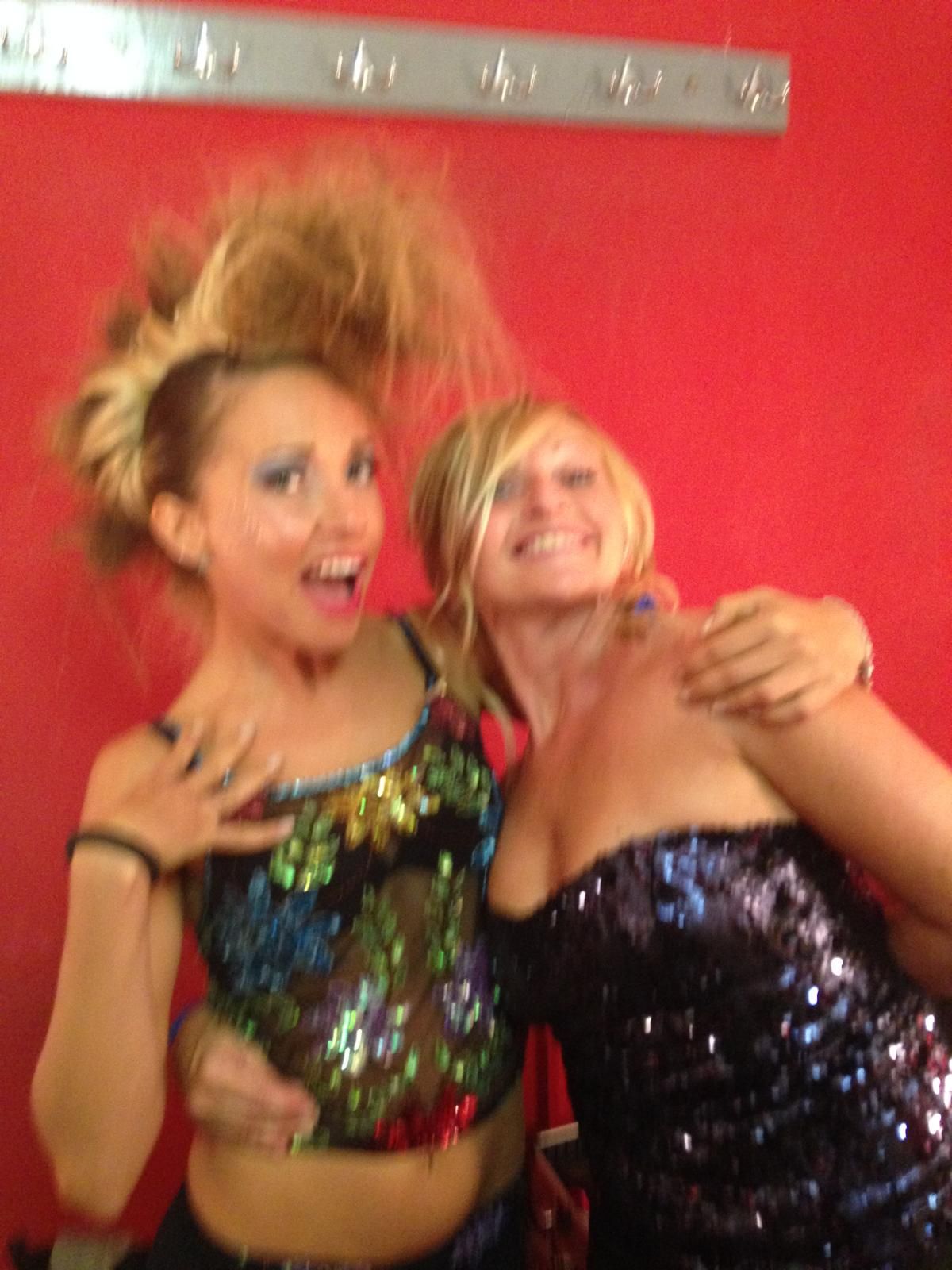 Due donne con top di paillettes, abbracciate. Una con i capelli biondi e selvaggi, l'altra con un abito senza spalline; sfondo rosso.