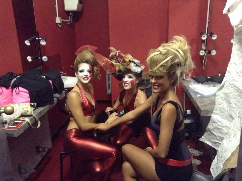 Tre artisti in costume nel backstage: trucco, abiti rossi/neri, capelli elaborati, posano insieme.