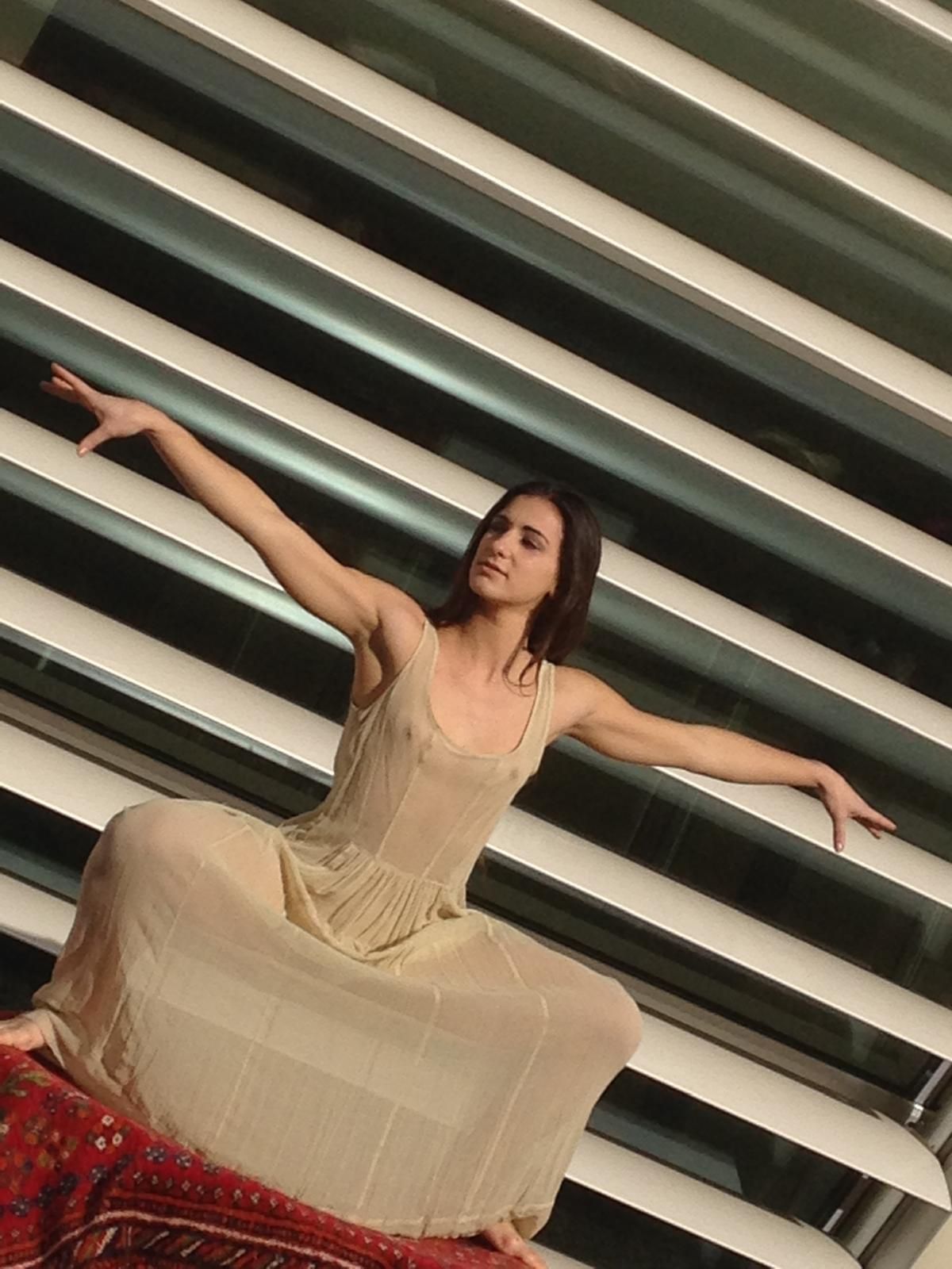 Donna in abito leggero, braccia tese, seduta su un tappeto a motivi geometrici su sfondo a righe.