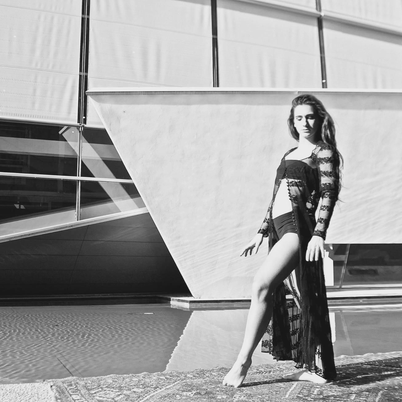 Donna in lingerie nera e vestaglia posa a bordo piscina. Edificio moderno sullo sfondo. Foto in bianco e nero.