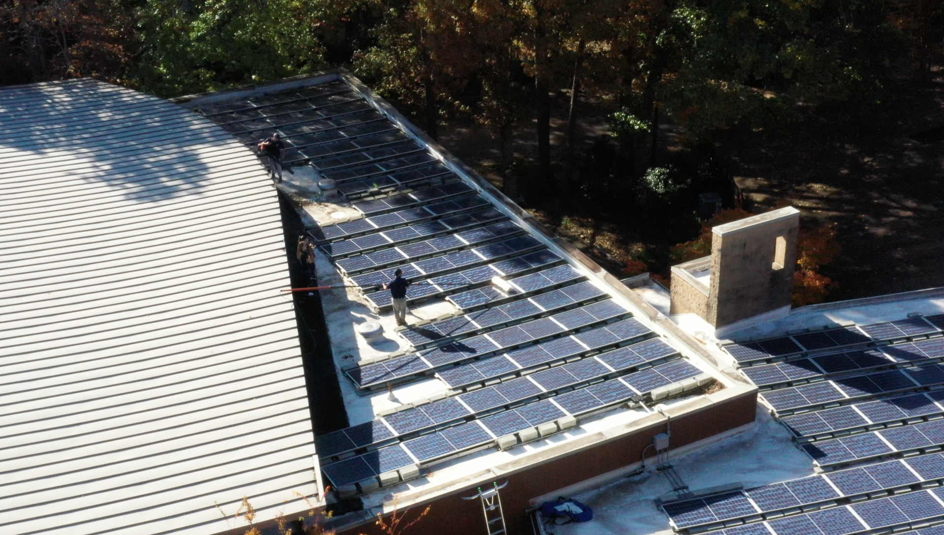 An aerial view shows workers cleaning rows of solar panels on a flat commercial roof 