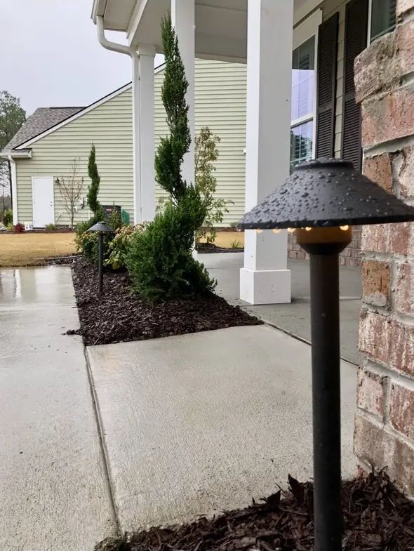 Path lined with shrubs and dark landscape lights, leading to a home's porch under a cloudy sky.