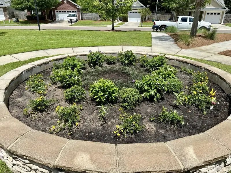 Circular flower bed with shrubs and dark soil, surrounded by a stone border. Houses and a truck are in the background.