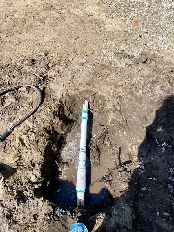 A dug-out hole reveals a white PVC pipe wrapped in white tape, with water pooling at the bottom.