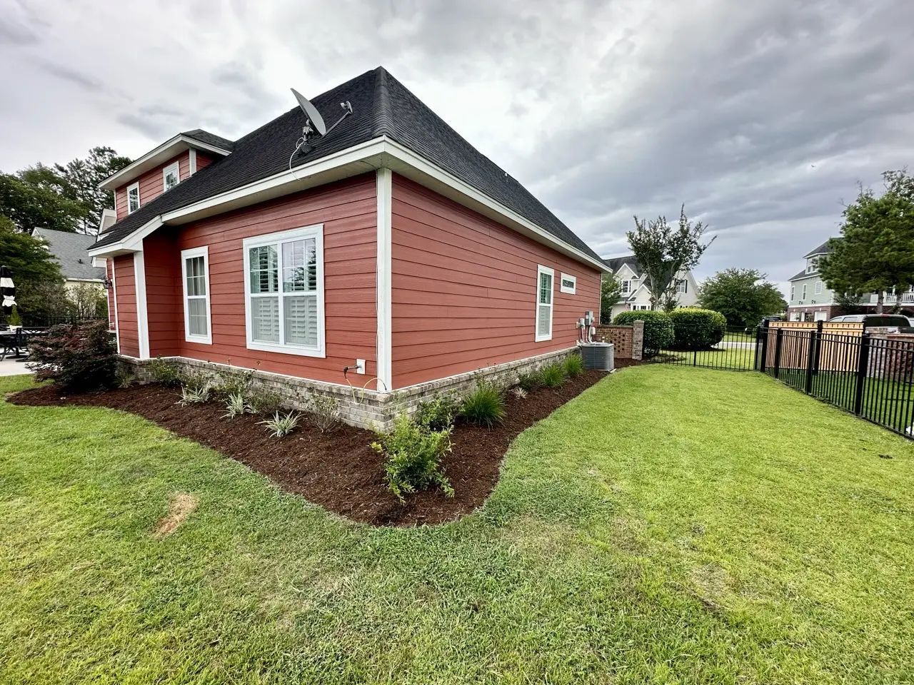 Red house with dark roof, white trim, and a well-manicured lawn with mulch and shrubs under a cloudy sky.