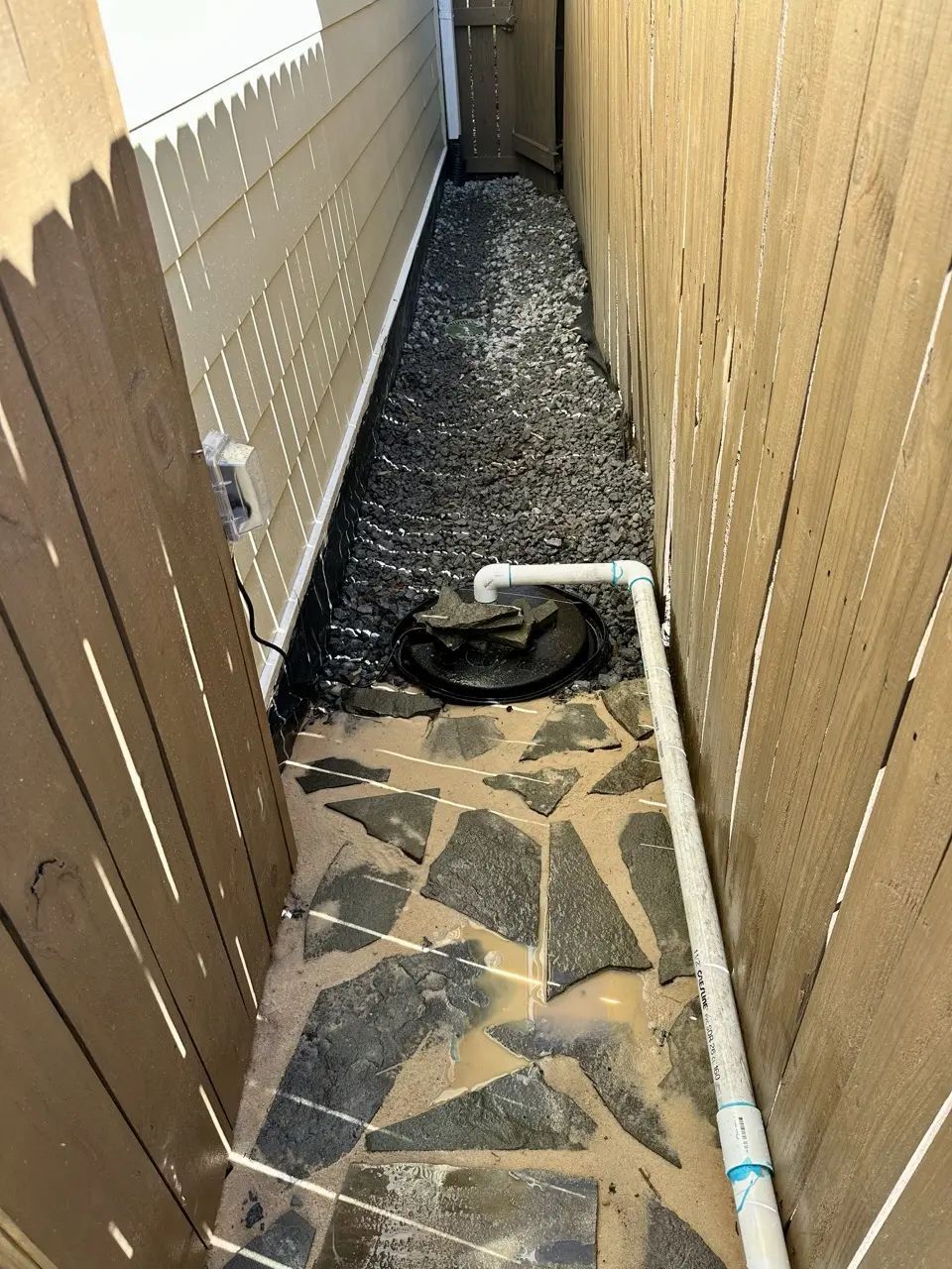 Narrow walkway between two wooden fences, with dark gravel and a stone pathway, leading to a sump pump.
