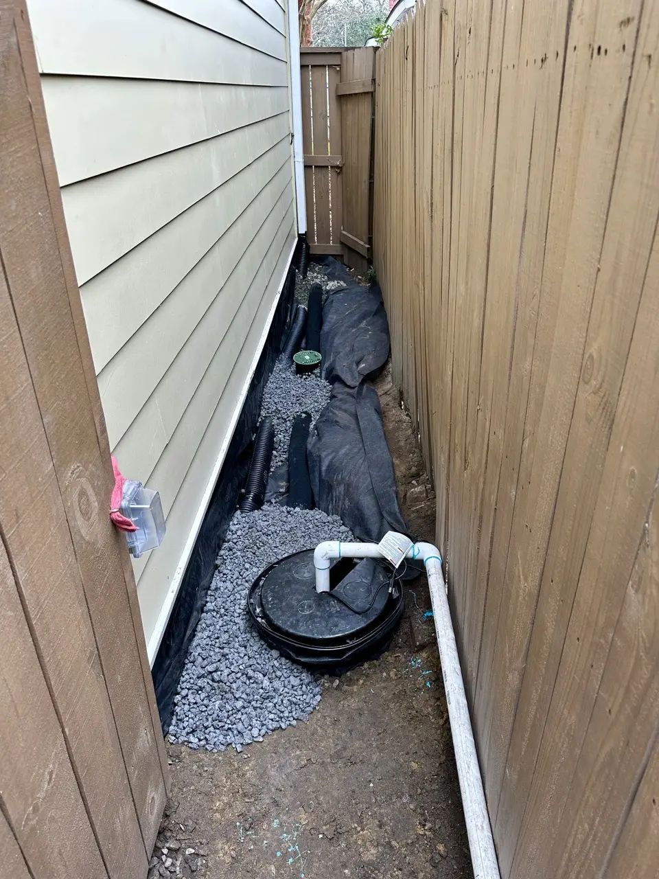 Narrow alley between tan house and wooden fence, with gravel, drainage system and pipe.