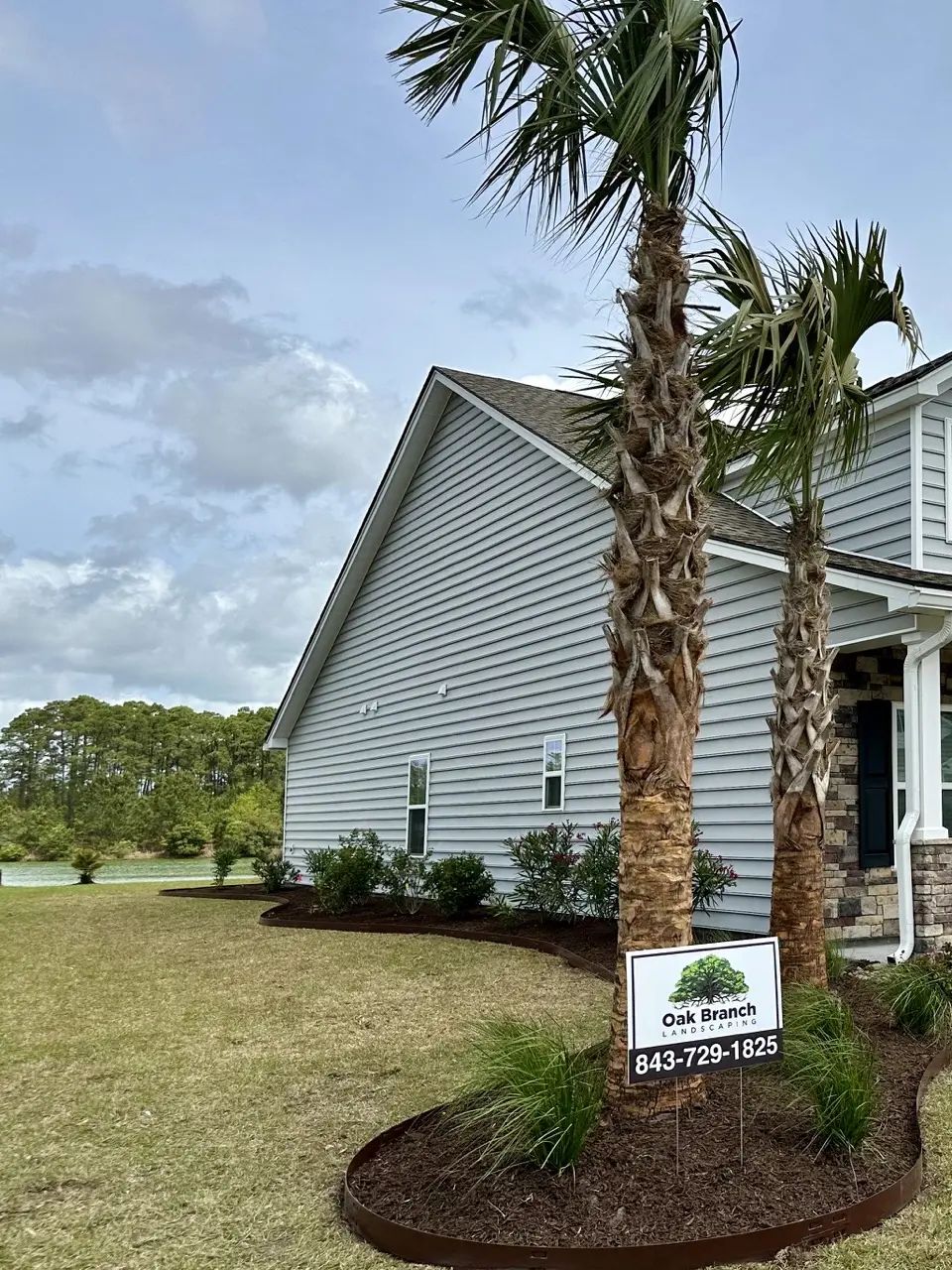 Two palm trees in front of a light blue house. Landscaping with dark mulch and grass in front. Cloudy sky.