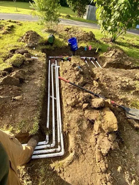 Trenches with white pipes being installed in a grassy area, a blue bucket sits nearby.