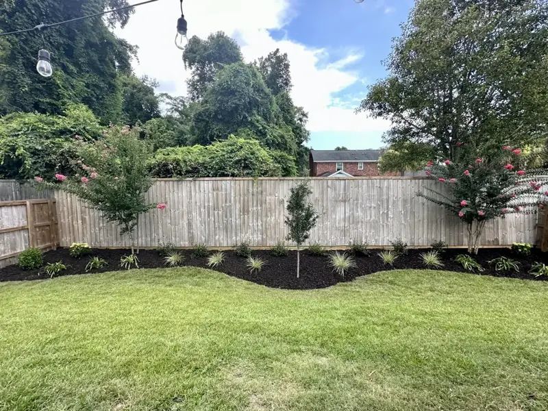 Backyard with a wooden fence, green lawn, flowerbed with dark mulch, and trees under a partly cloudy sky.