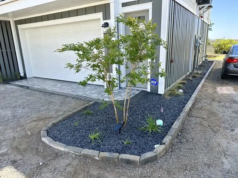 Curb-edged bed with black gravel, young tree, and small plants next to a garage and house. Gray siding, paving.
