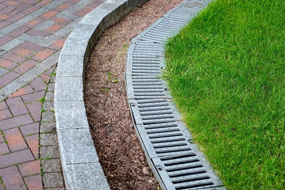 Curved brick walkway bordered by a gray stone curb and a metal drainage channel next to a grassy lawn.