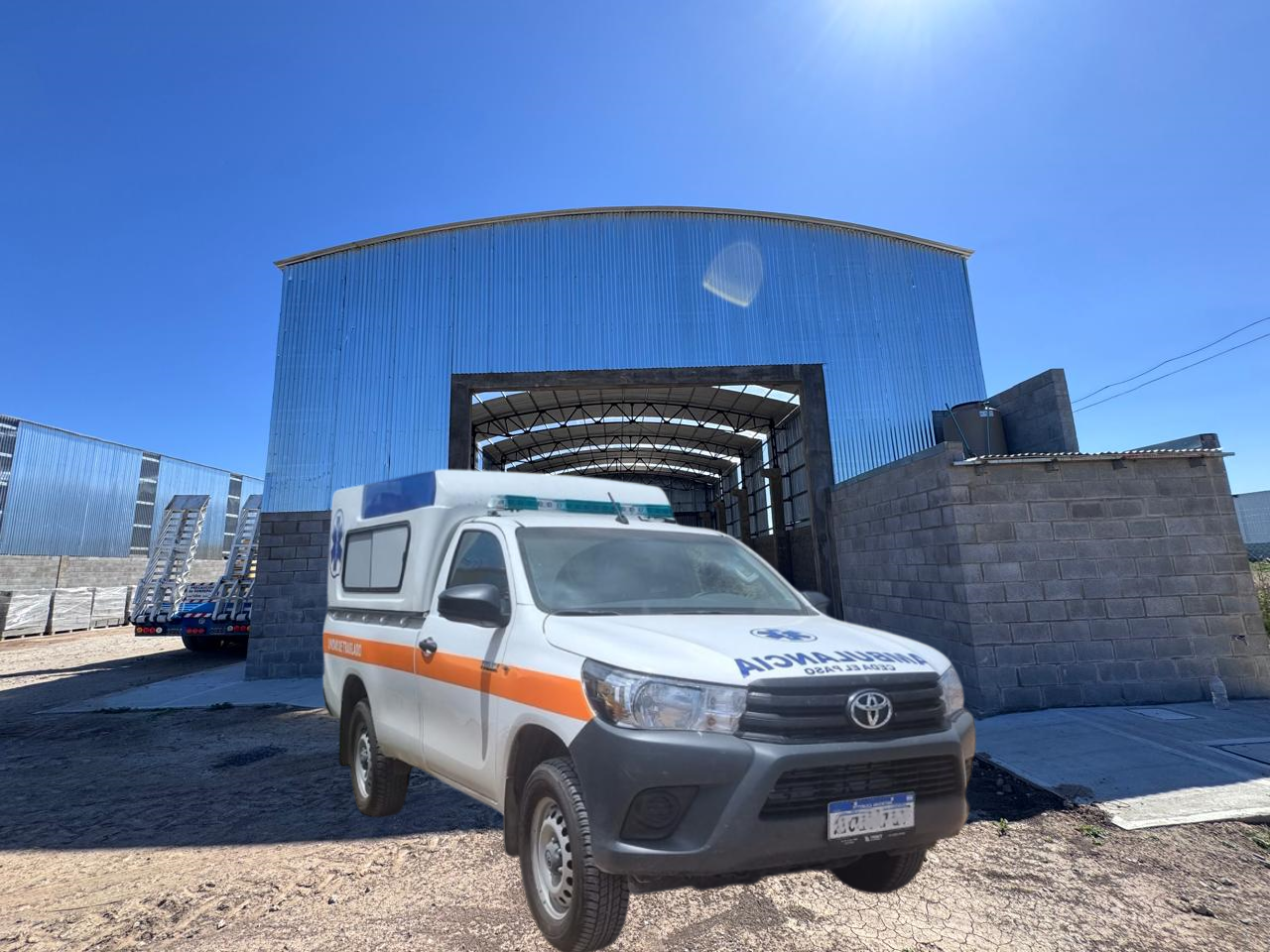 Camión ambulancia blanco con rayas naranjas estacionado frente a un gran edificio de metal bajo un cielo azul claro.