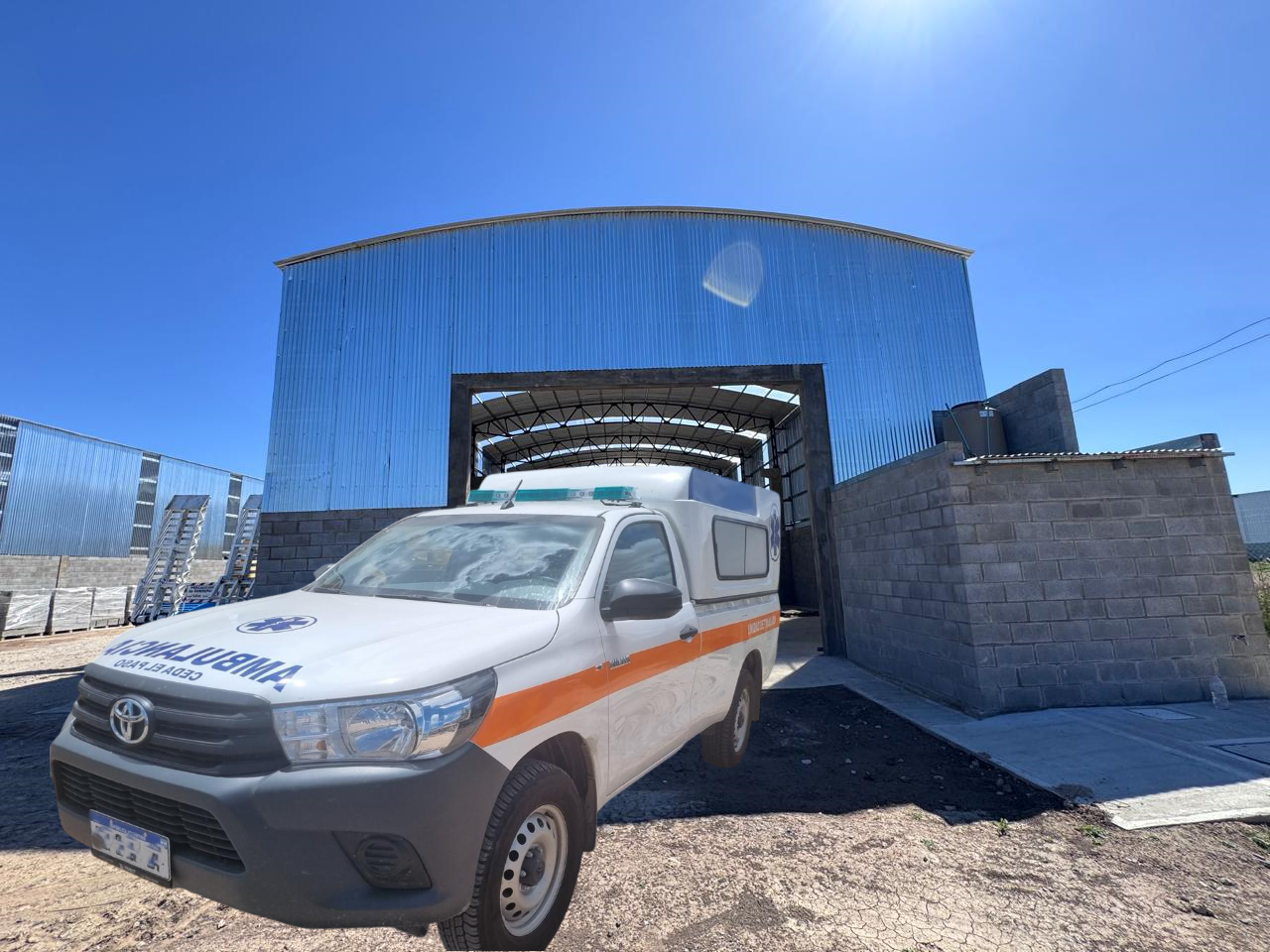 Ambulancia blanca estacionada frente a un edificio metálico azul con la entrada abierta. Día soleado.