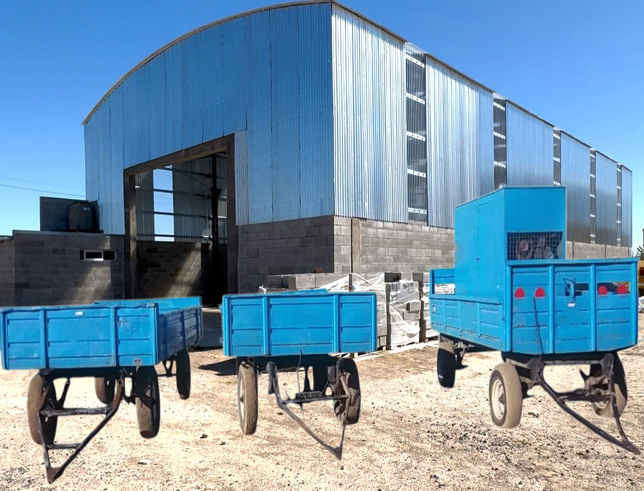 Tres carros agrícolas azules frente a un almacén de metal bajo un cielo despejado.