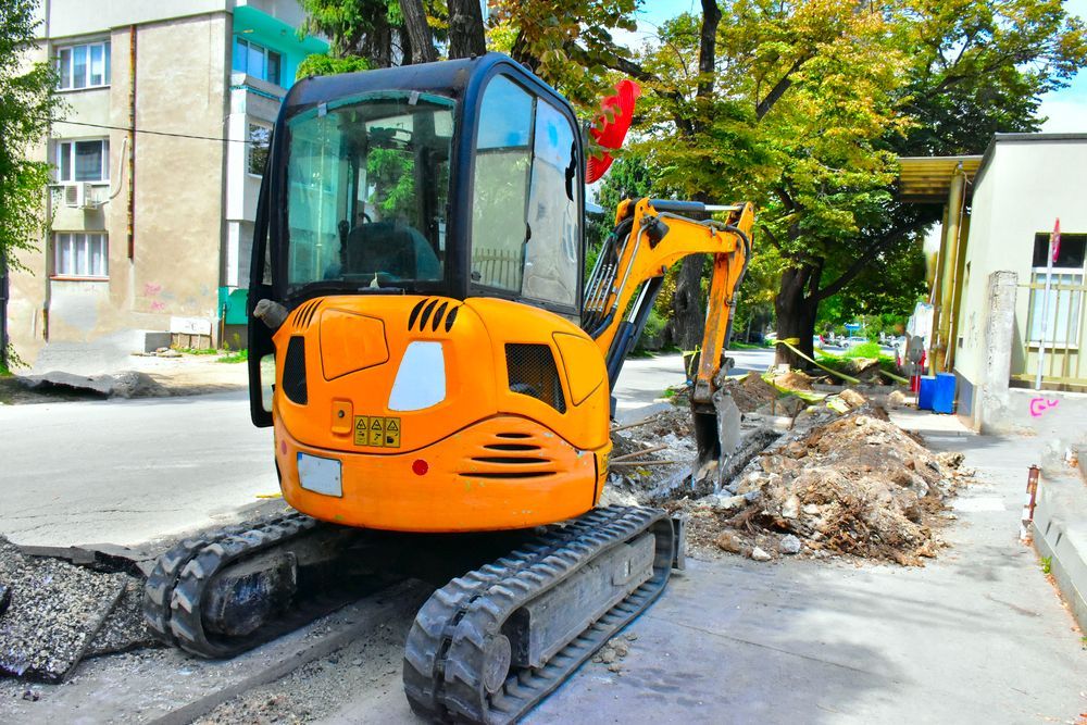 Excavadora naranja en una acera, cavando cerca de un edificio y un árbol.