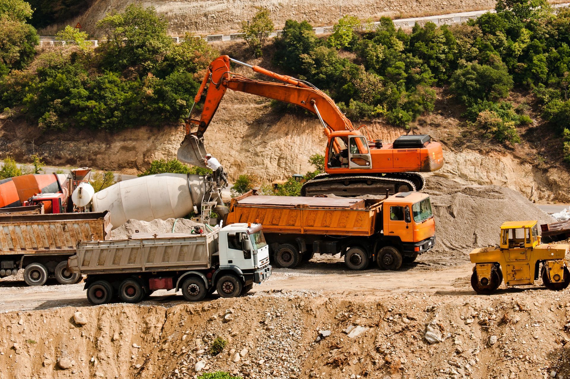 Sitio de construcción con excavadora cargando un camión volquete; hay otros camiones y maquinaria presentes.