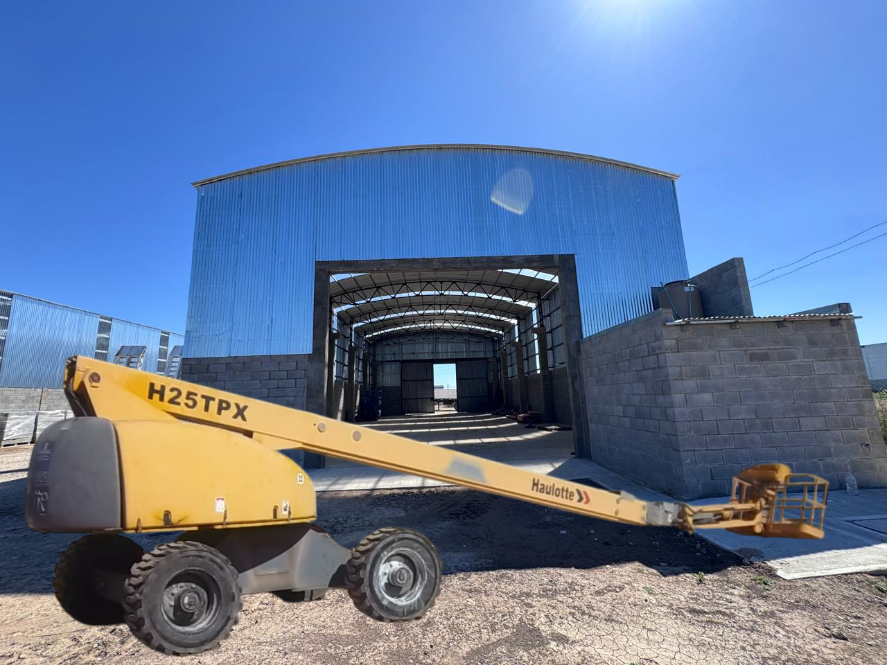 Plataforma elevadora amarilla frente a un edificio metálico parcialmente construido. Cielo azul.