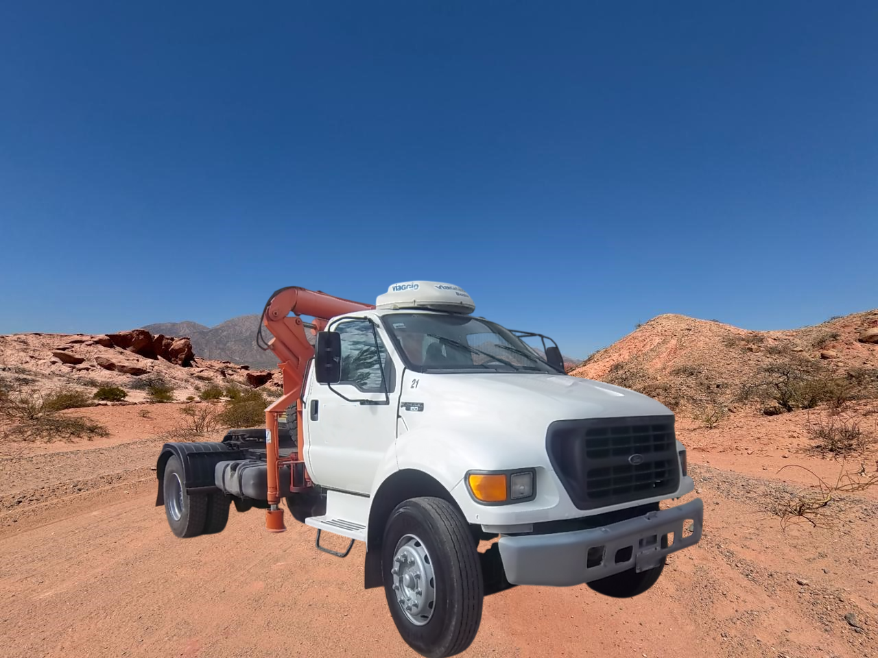 Camión grúa blanco en un camino de tierra en un paisaje desértico con rocas de color marrón rojizo y cielo azul.