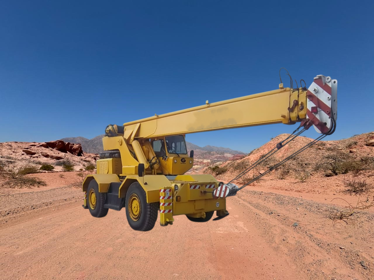 Grúa móvil amarilla en un camino de tierra en un paisaje desértico con formaciones rocosas rojas. Cielo azul de fondo.