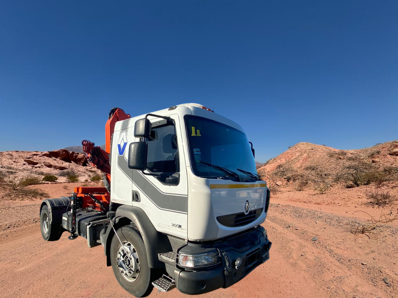 Camión blanco con una grúa naranja en un camino de tierra contra un cielo azul y un paisaje desértico.