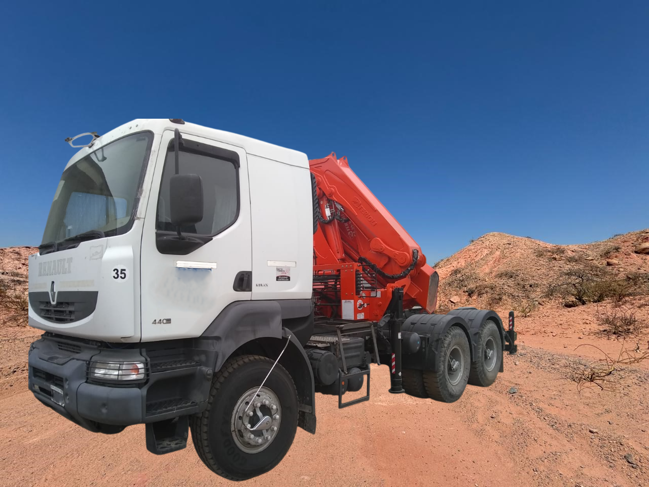 Camión blanco con grúa naranja en un camino de tierra contra un cielo azul, montañas al fondo.