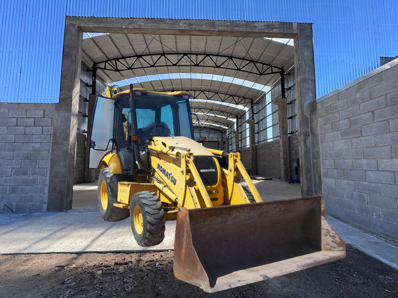 Retroexcavadora Komatsu amarilla dentro de un edificio de hormigón, bajo un techo de metal, con un conductor en la cabina.