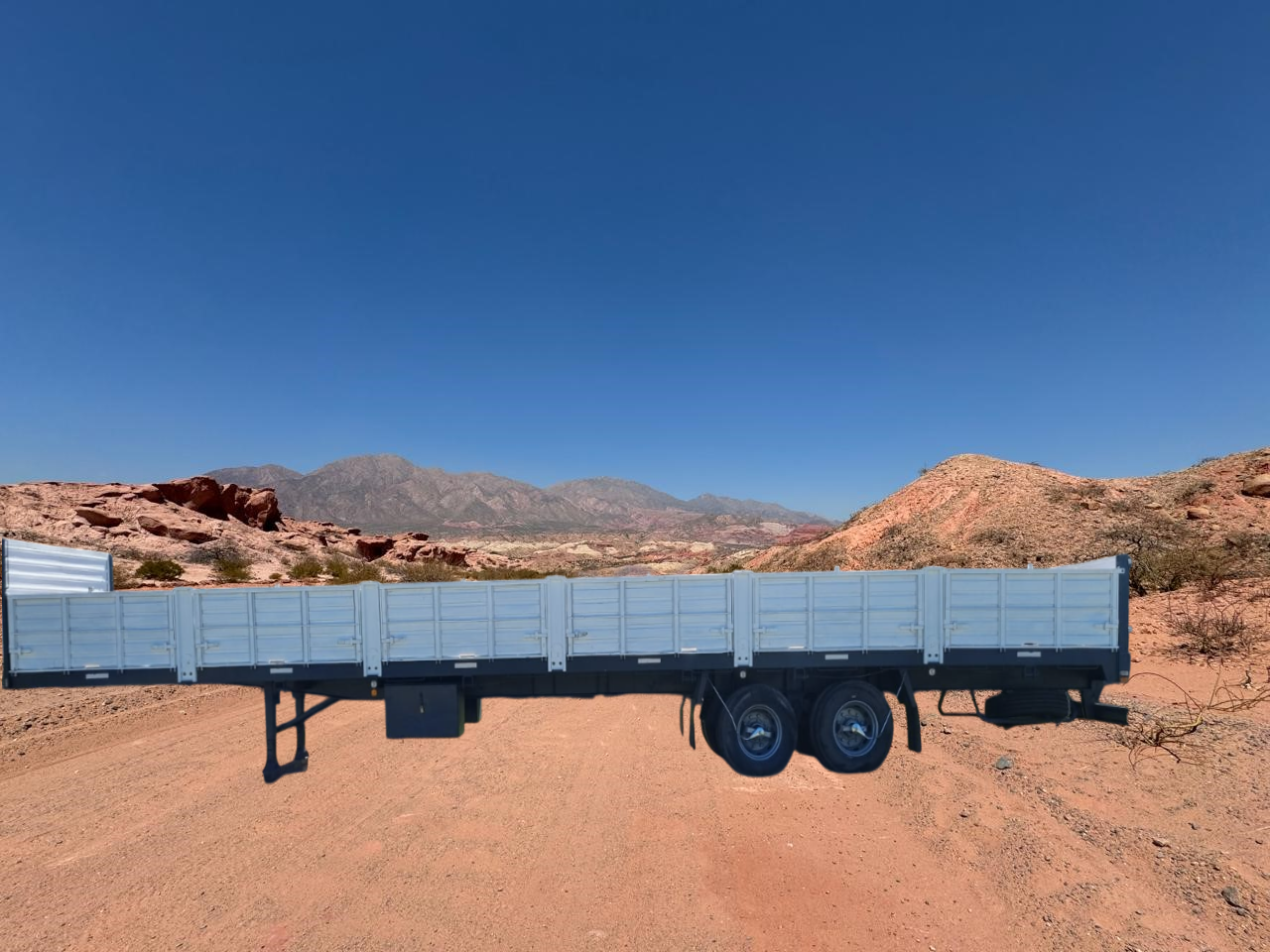 Remolque de plataforma en un camino de tierra en un paisaje desértico con montañas al fondo bajo un cielo azul.