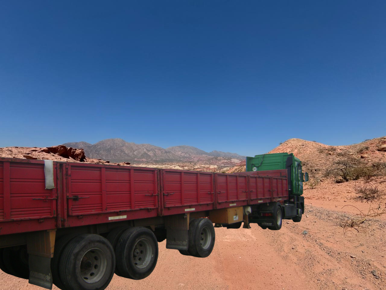 Camión de plataforma roja en camino de tierra, montañas al fondo, cielo azul.