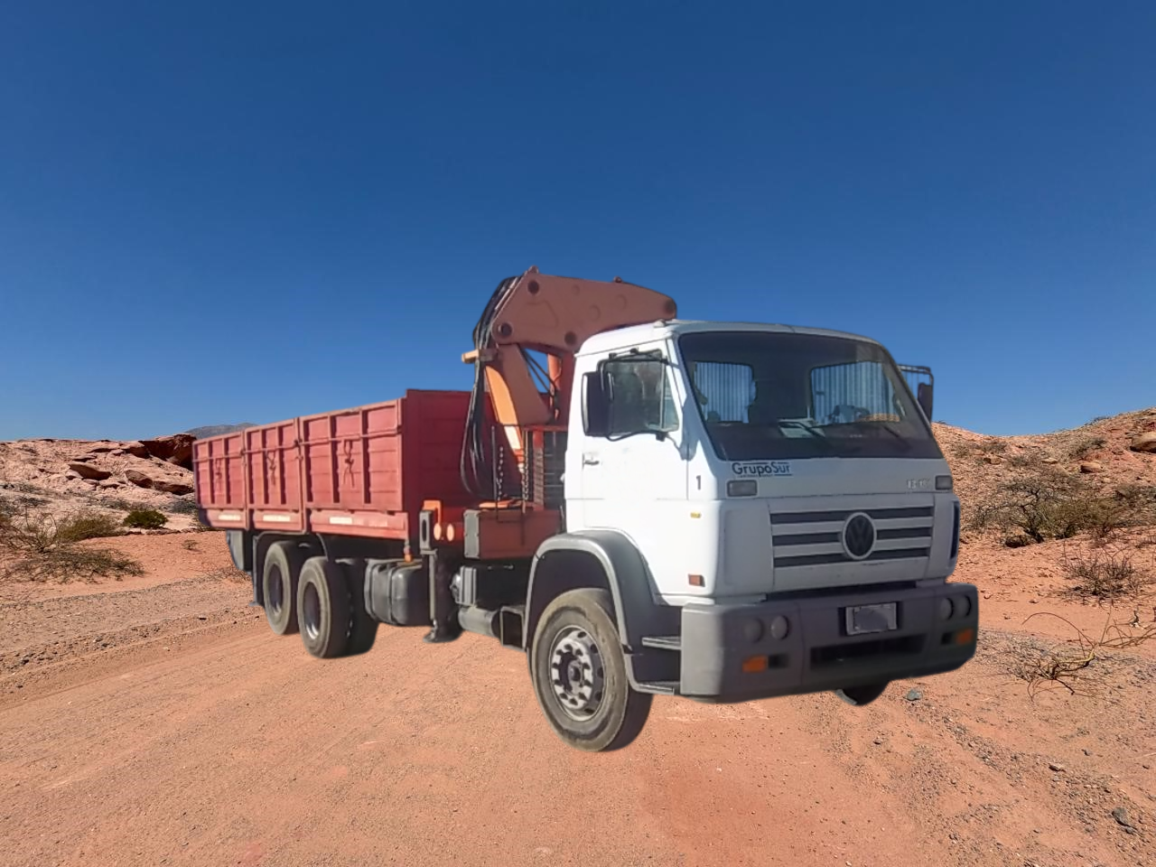 Un camión grúa blanco en un camino de tierra en un paisaje desértico con acantilados rojos bajo un cielo azul.