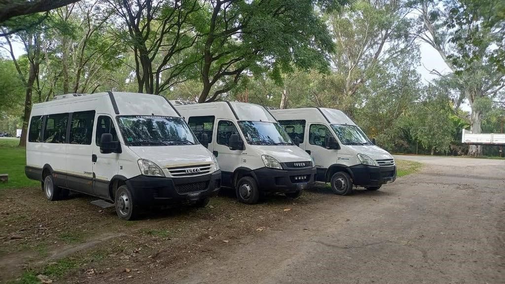 Tres furgonetas blancas estacionadas en una zona de grava, bajo árboles.