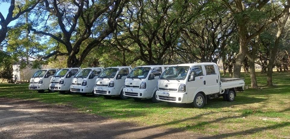 Siete camionetas Kia Bongo blancas estacionadas en fila sobre el césped bajo los árboles.