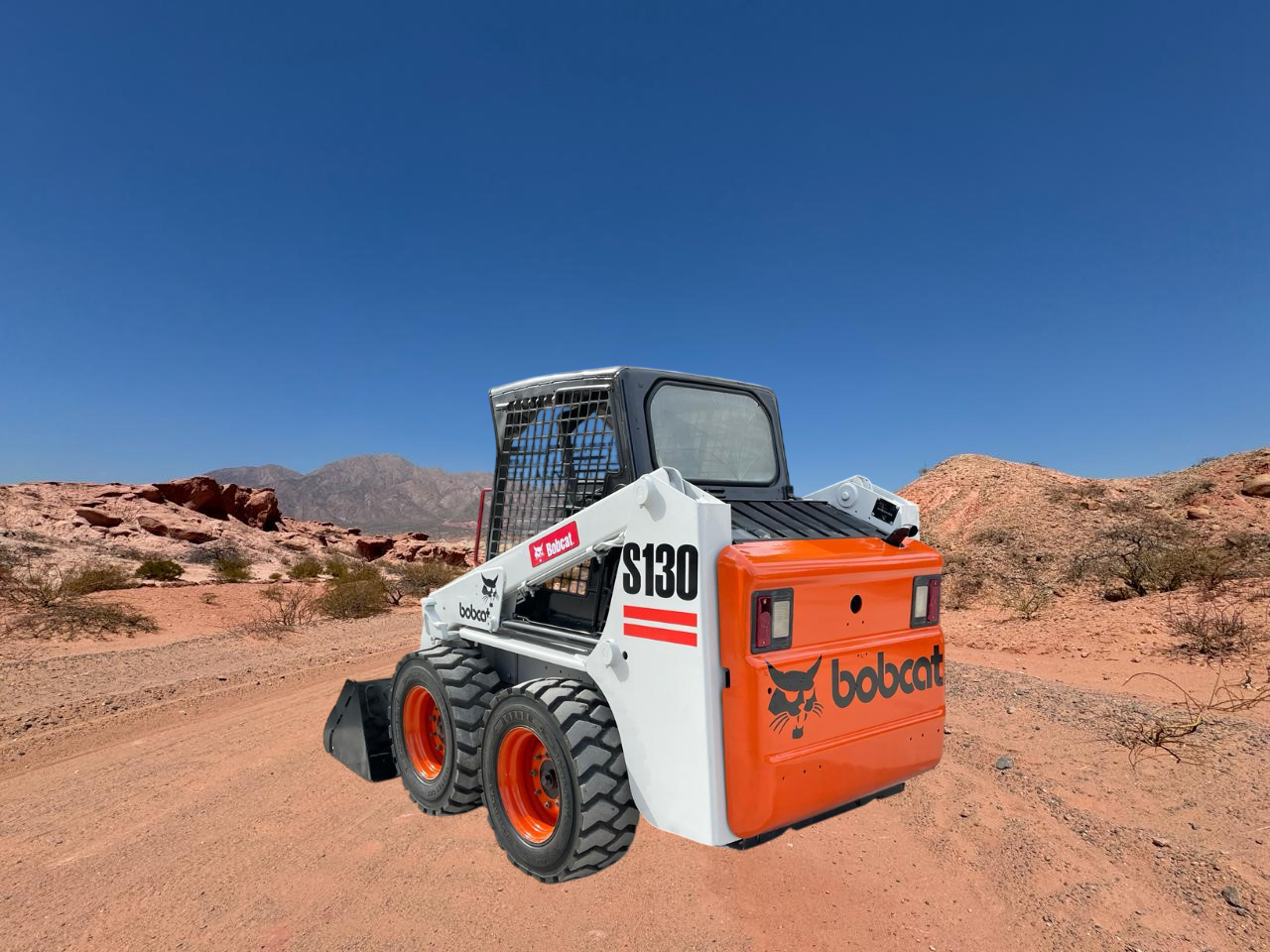 Cargadora compacta Bobcat S130 en un camino de tierra en un paisaje desértico con formaciones rocosas rojas y un cielo azul.