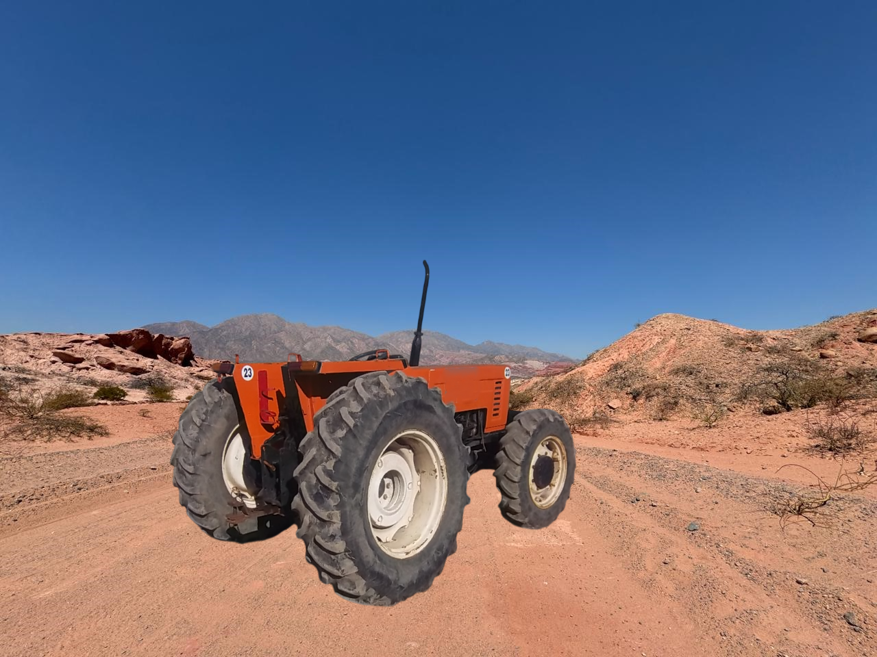 Tractor naranja en un camino de tierra en un paisaje desértico con montañas y un cielo azul claro.