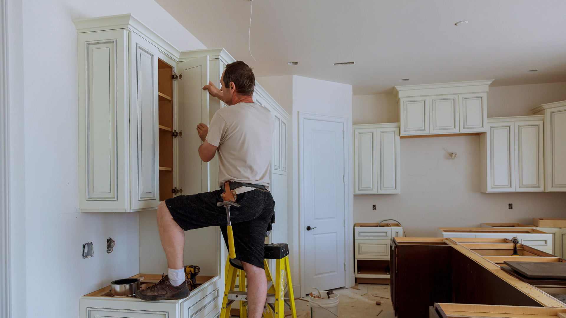 A man is standing on a ladder in a kitchen installing cabinets.