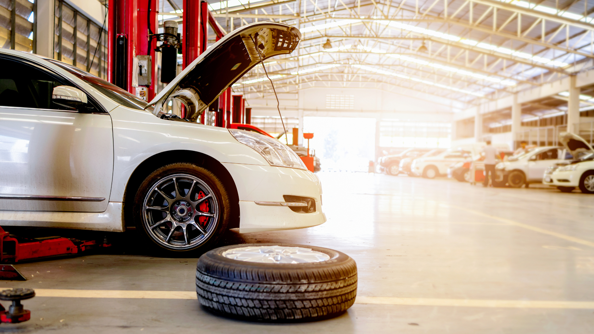 White car on a lift with hood open in a repair shop; tire on the floor.