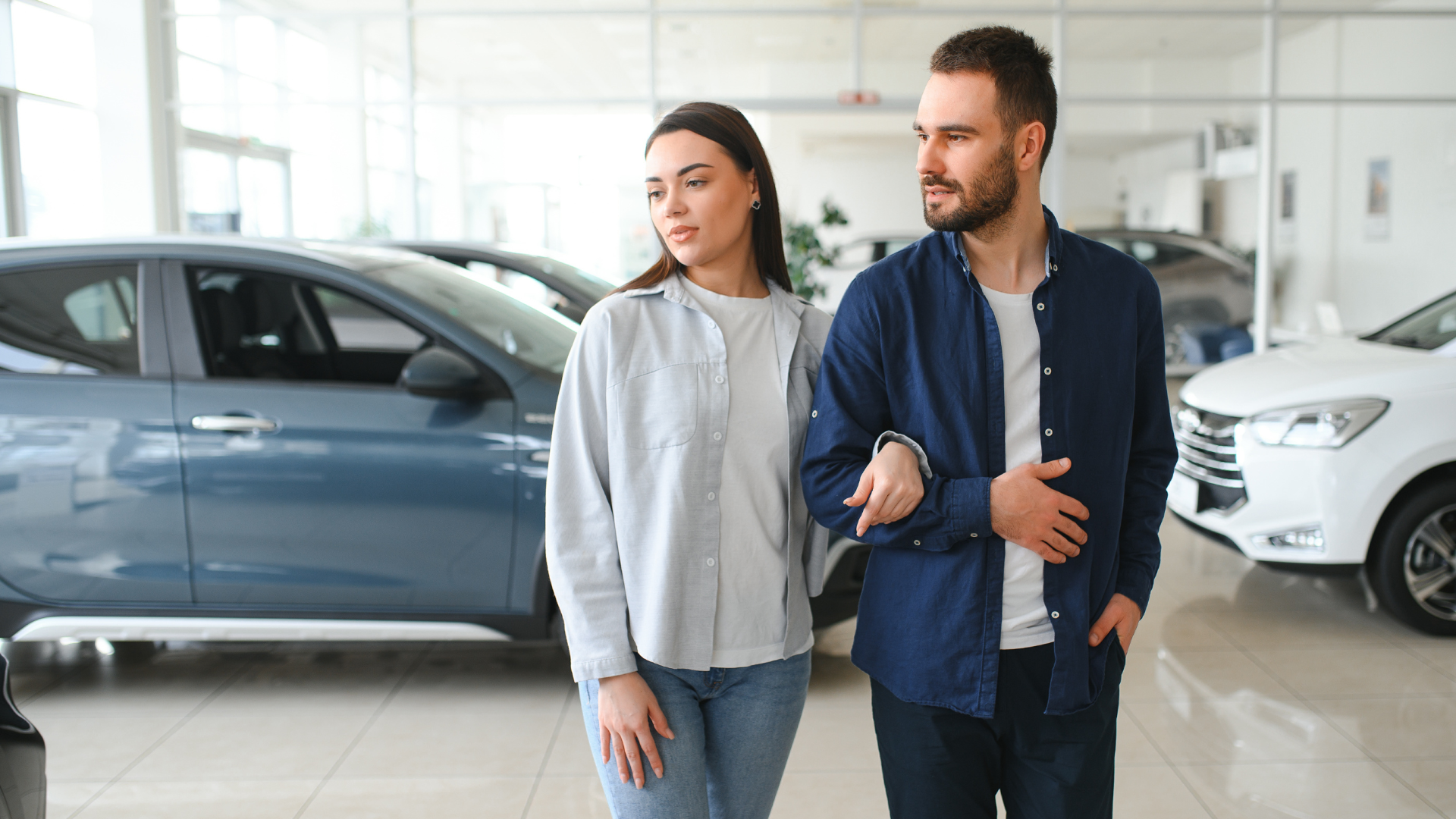 Couple looking at cars in a dealership showroom; man in blue, woman in light blue, cars in background.
