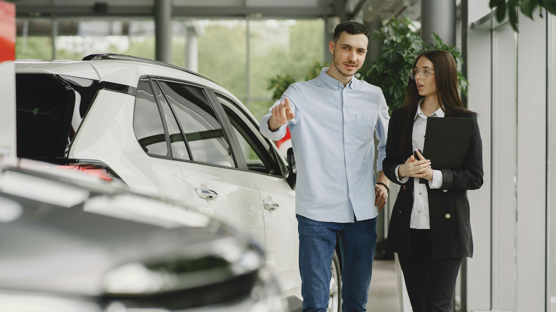 Man and woman examining a car in a showroom. The man gestures at the car as they walk.
