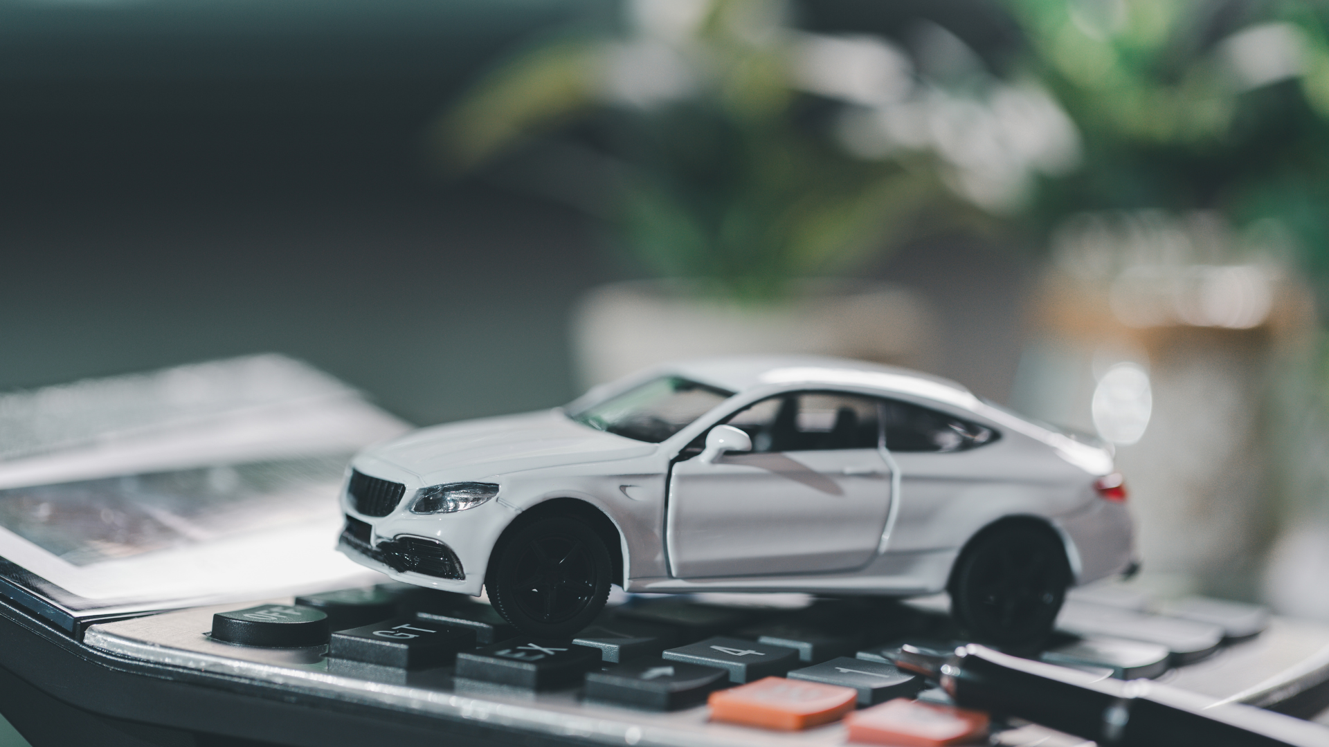 A white toy car on a calculator, near a pen and potted plant, suggesting cost analysis.