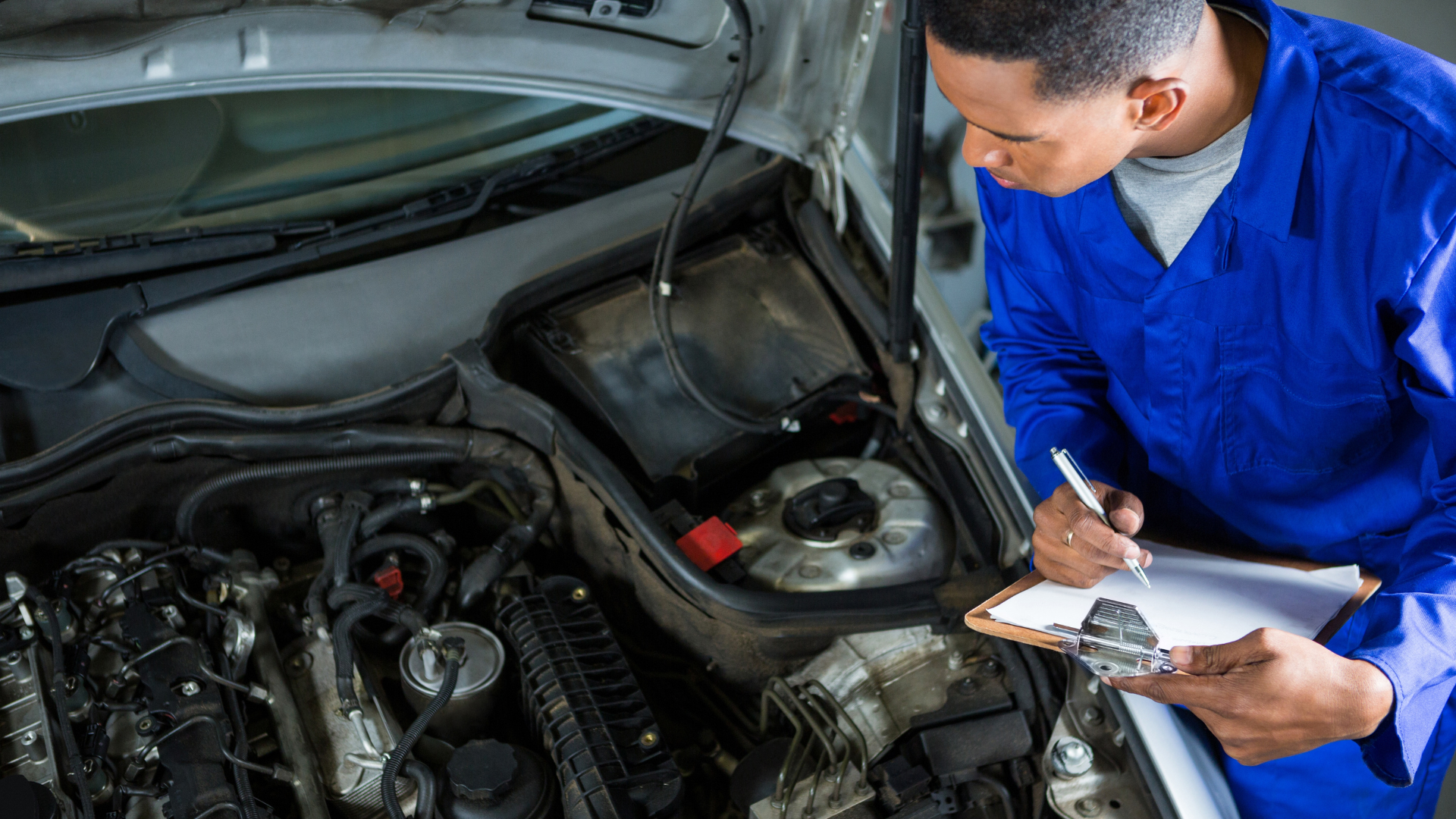 Mechanic in blue jumpsuit inspecting car engine, writing on clipboard.