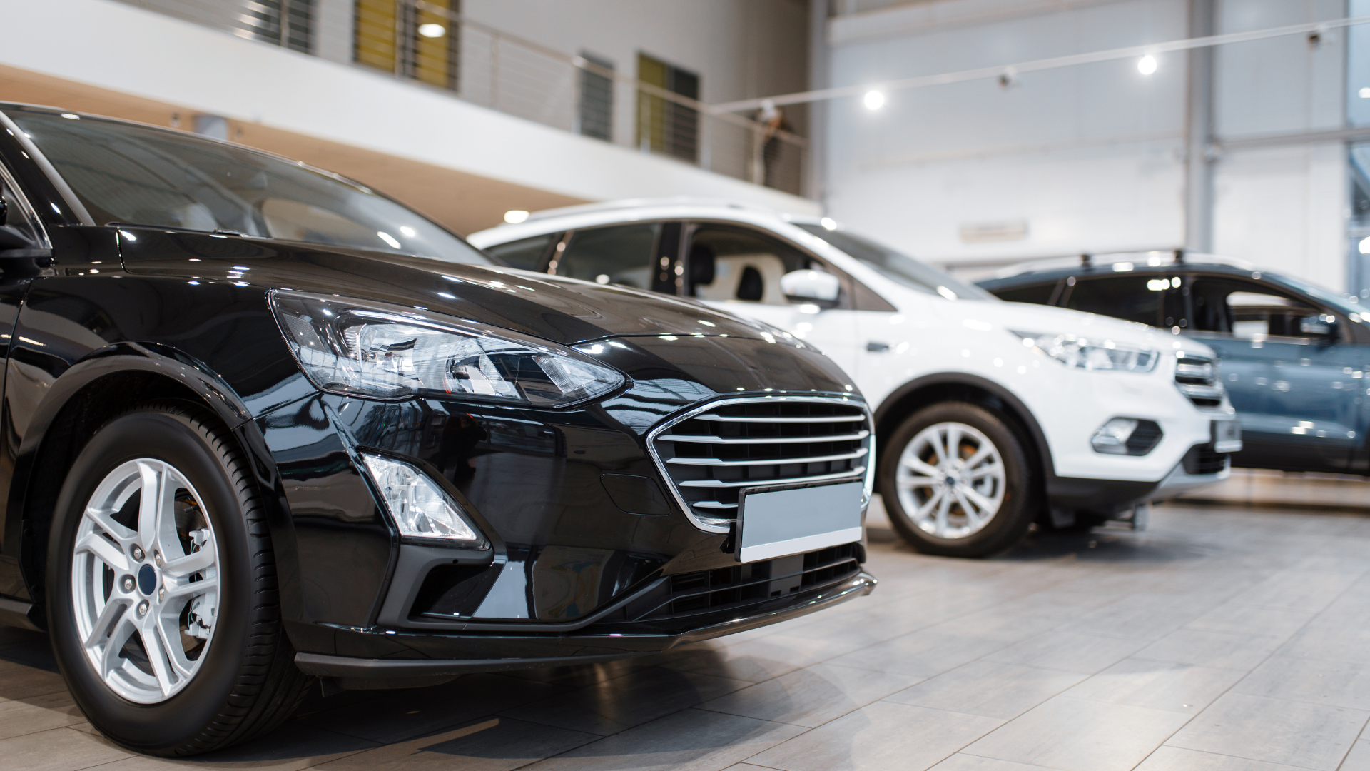 Cars displayed inside a car dealership showroom: black sedan in front, white SUV in the middle, and blue SUV in the back.