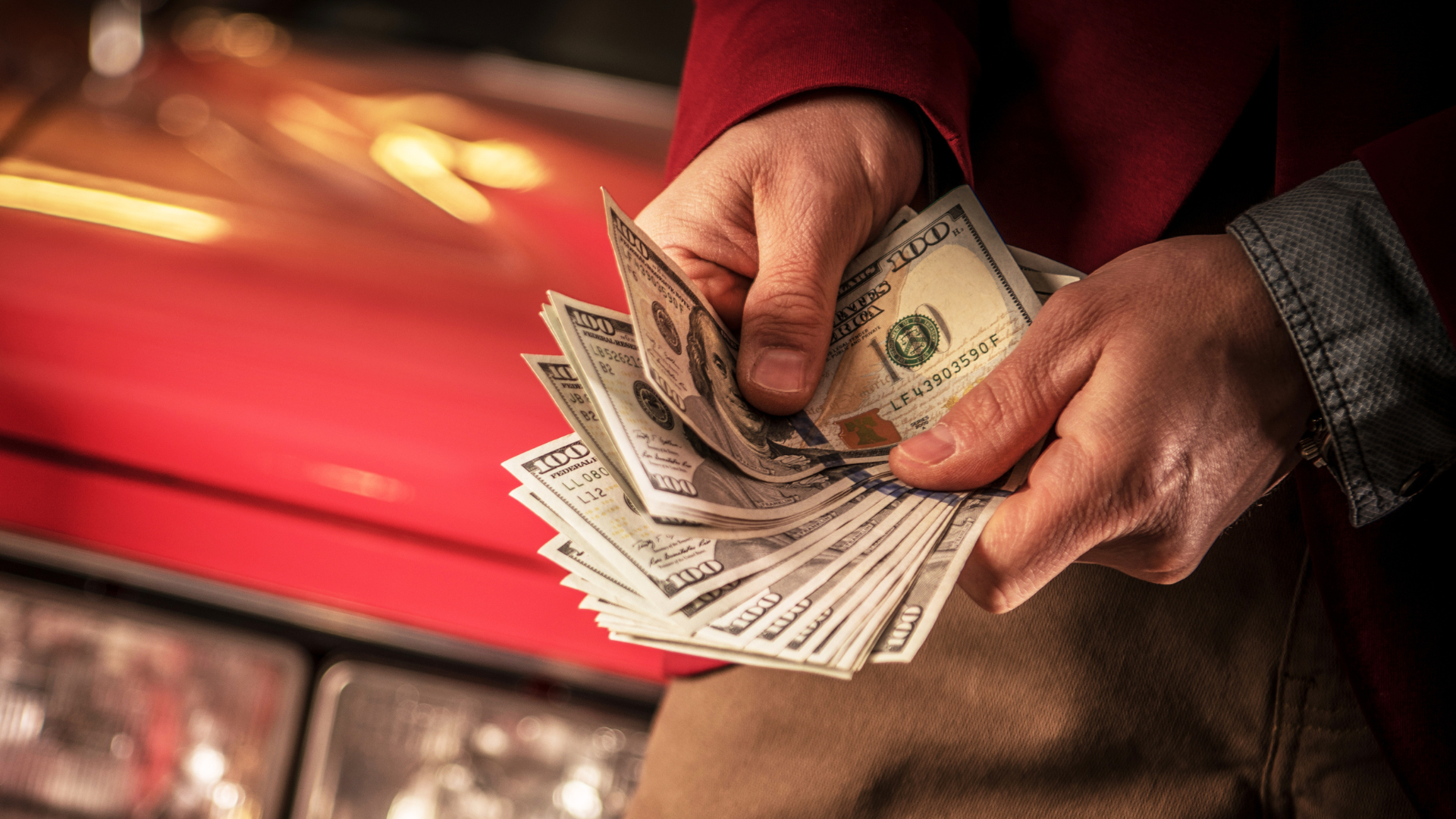 A person is holding a bunch of money in front of a red car.