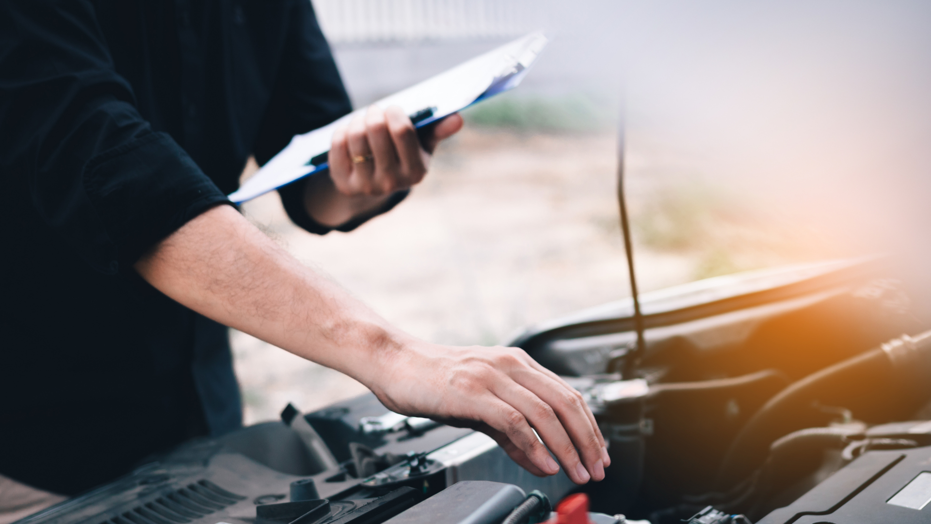 A man is looking under the hood of a car while holding a clipboard.