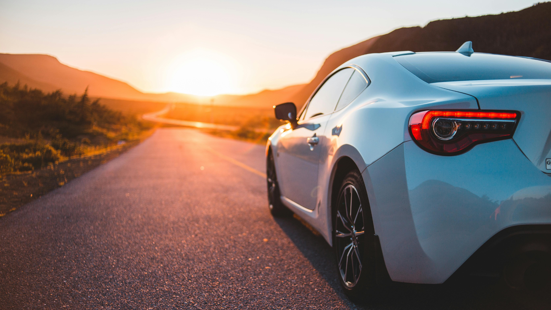 White sports car on a winding road at sunset, mountains in the background.