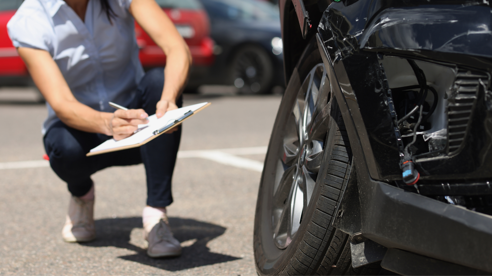 A woman is kneeling down in front of a damaged car and writing on a clipboard.