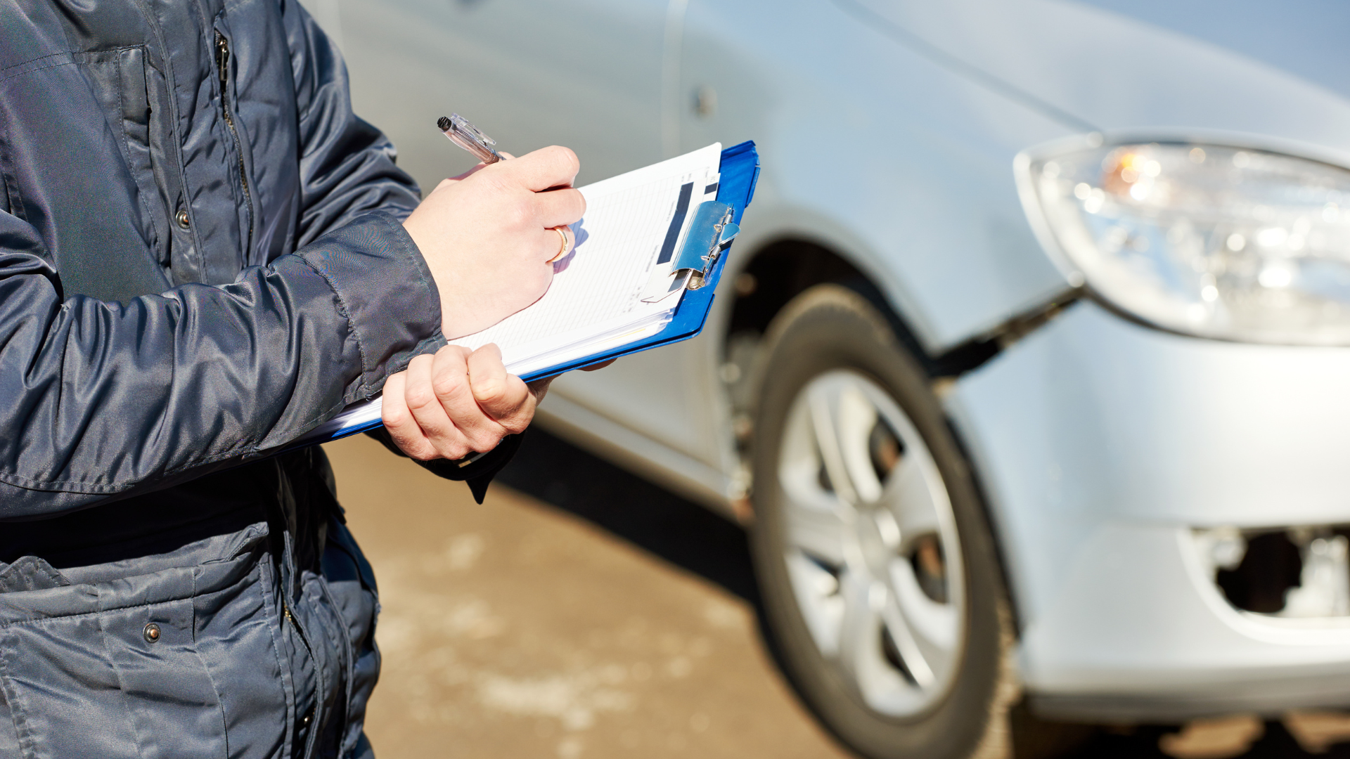 A man is writing on a clipboard in front of a damaged car.