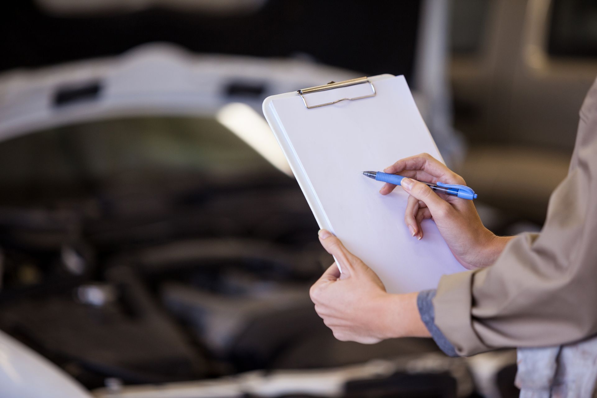 Mechanic inspecting car engine, writing on a clipboard with a pen.